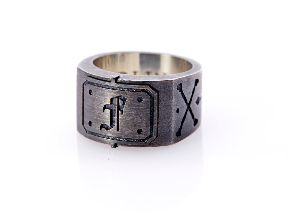 Oxidized sterling silver men’s signet ring with a thick flat band hand-crafted in New York. Semi-raised octagonal engraved top cap with a customizable insignia design of an engraved letter “F” in Old English Style lettering and 4 engraved dots at each corner. The initial on the ring is surrounded by a bold black engraved border that follows the inside edge of the rectangle. The rectangle’s top and bottom edge is accented by a small lip in the center that wraps around the corner edge of the ring.