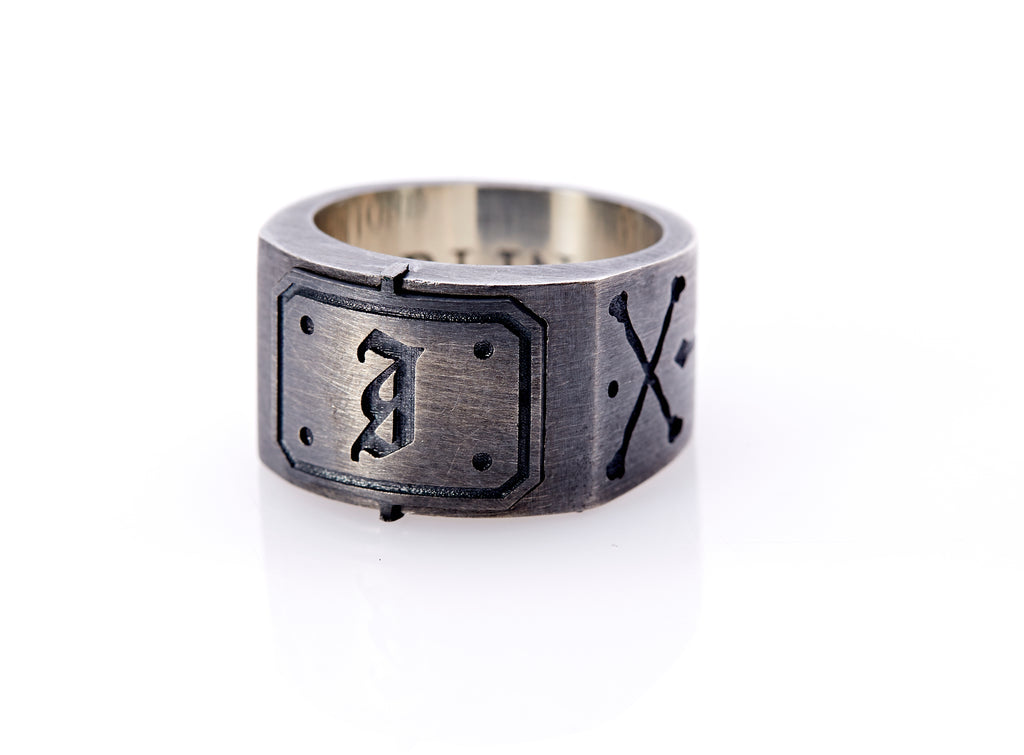 Oxidized sterling silver men’s signet ring with a thick flat band hand-crafted in New York. Semi-raised octagonal engraved top cap with a customizable insignia design of an engraved letter “I” in Old English Style lettering and 4 engraved dots at each corner. The initial on the ring is surrounded by a bold black engraved border that follows the inside edge of the rectangle. The rectangle’s top and bottom edge is accented by a small lip in the center that wraps around the corner edge of the ring.