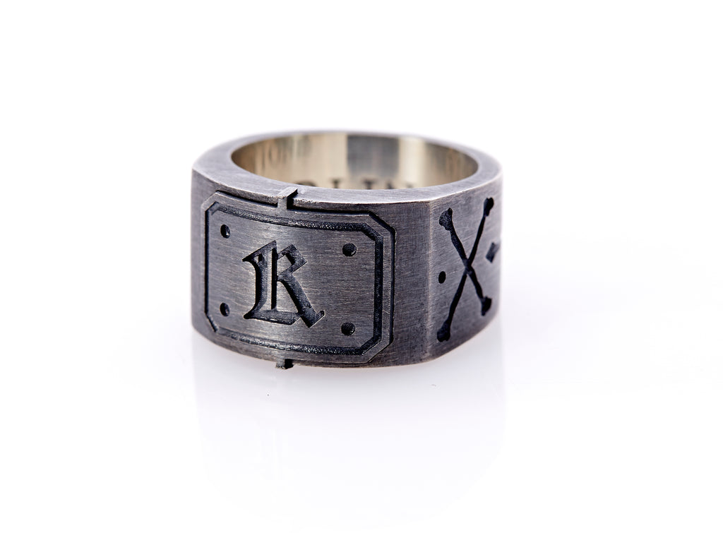 Oxidized sterling silver men’s signet ring with a thick flat band hand-crafted in New York. Semi-raised octagonal engraved top cap with a customizable insignia design of an engraved letter “K” in Old English Style lettering and 4 engraved dots at each corner. The initial on the ring is surrounded by a bold black engraved border that follows the inside edge of the rectangle. The rectangle’s top and bottom edge is accented by a small lip in the center that wraps around the corner edge of the ring.