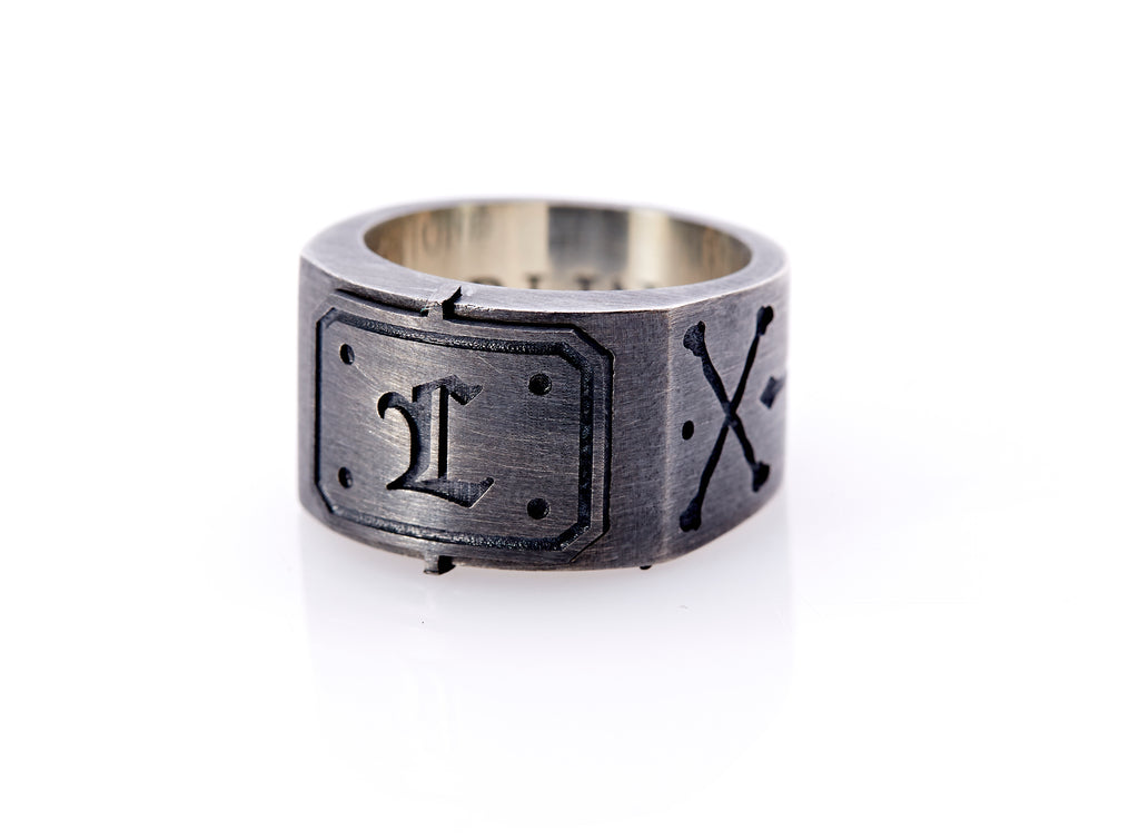 Oxidized sterling silver men’s signet ring with a thick flat band hand-crafted in New York. Semi-raised octagonal engraved top cap with a customizable insignia design of an engraved letter “L” in Old English Style lettering and 4 engraved dots at each corner. The initial on the ring is surrounded by a bold black engraved border that follows the inside edge of the rectangle. The rectangle’s top and bottom edge is accented by a small lip in the center that wraps around the corner edge of the ring.