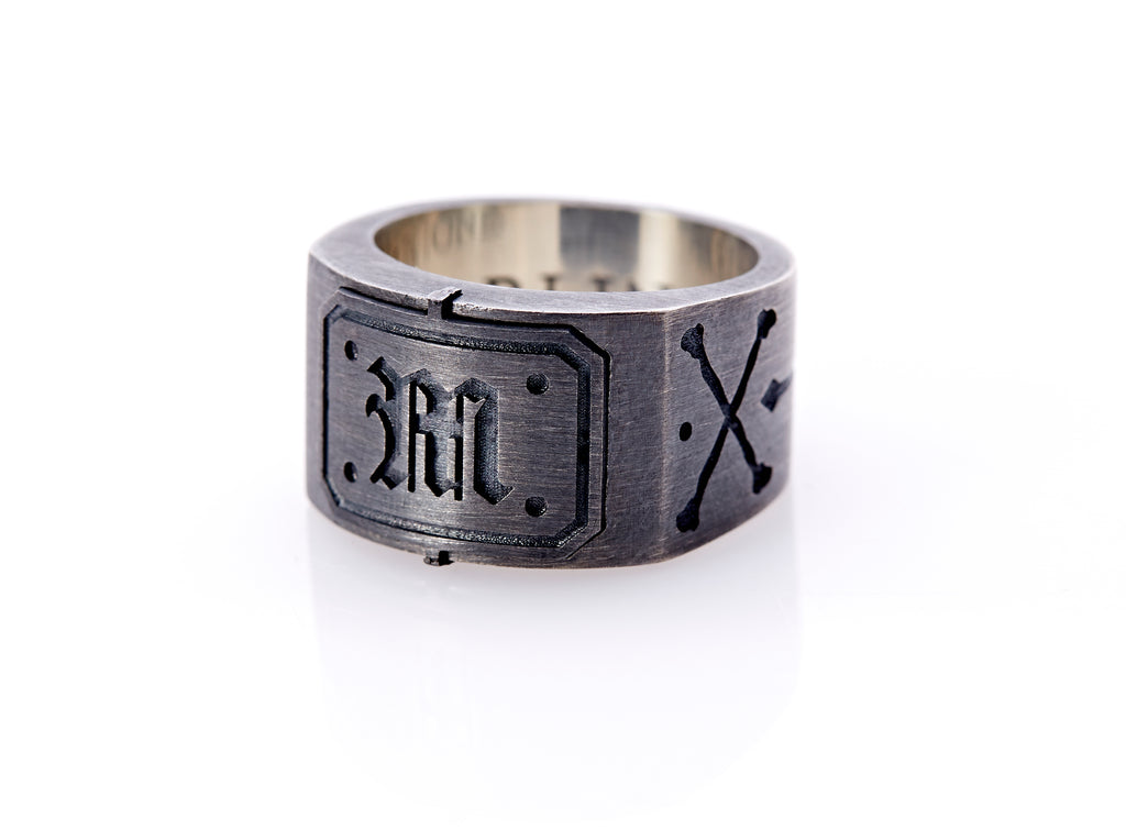 Oxidized sterling silver men’s signet ring with a thick flat band hand-crafted in New York. Semi-raised octagonal engraved top cap with a customizable insignia design of an engraved letter “M” in Old English Style lettering and 4 engraved dots at each corner. The initial on the ring is surrounded by a bold black engraved border that follows the inside edge of the rectangle. The rectangle’s top and bottom edge is accented by a small lip in the center that wraps around the corner edge of the ring.
