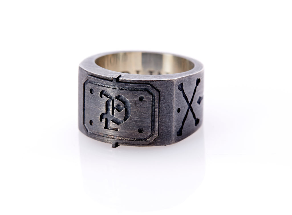 Oxidized sterling silver men’s signet ring with a thick flat band hand-crafted in New York. Semi-raised octagonal engraved top cap with a customizable insignia design of an engraved letter “P” in Old English Style lettering and 4 engraved dots at each corner. The initial on the ring is surrounded by a bold black engraved border that follows the inside edge of the rectangle. The rectangle’s top and bottom edge is accented by a small lip in the center that wraps around the corner edge of the ring.