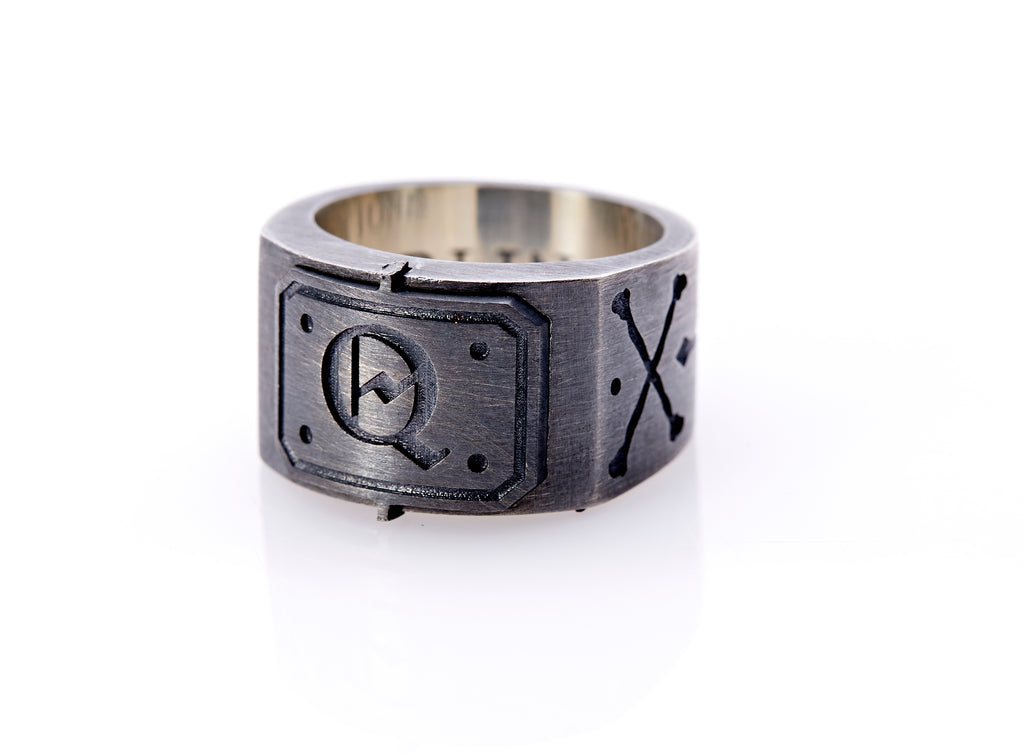 Oxidized sterling silver men’s signet ring with a thick flat band hand-crafted in New York. Semi-raised octagonal engraved top cap with a customizable insignia design of an engraved letter “Q” in Old English Style lettering and 4 engraved dots at each corner. The initial on the ring is surrounded by a bold black engraved border that follows the inside edge of the rectangle. The rectangle’s top and bottom edge is accented by a small lip in the center that wraps around the corner edge of the ring.