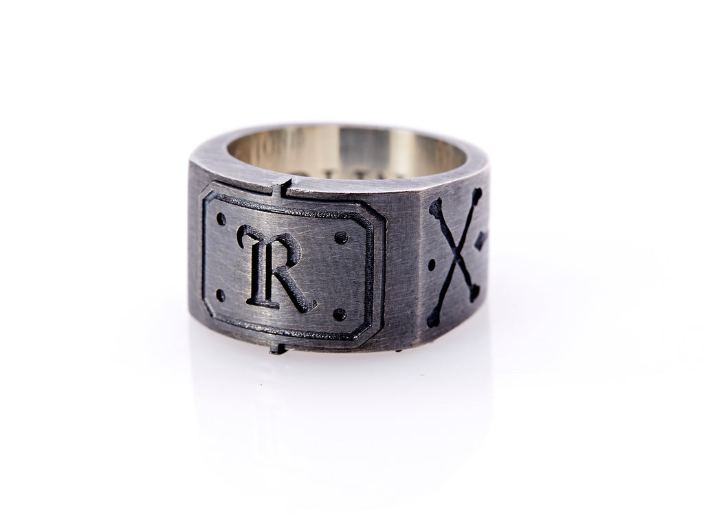 Oxidized sterling silver men’s signet ring with a thick flat band hand-crafted in New York. Semi-raised octagonal engraved top cap with a customizable insignia design of an engraved letter “R” in Old English Style lettering and 4 engraved dots at each corner. The initial on the ring is surrounded by a bold black engraved border that follows the inside edge of the rectangle. The rectangle’s top and bottom edge is accented by a small lip in the center that wraps around the corner edge of the ring.