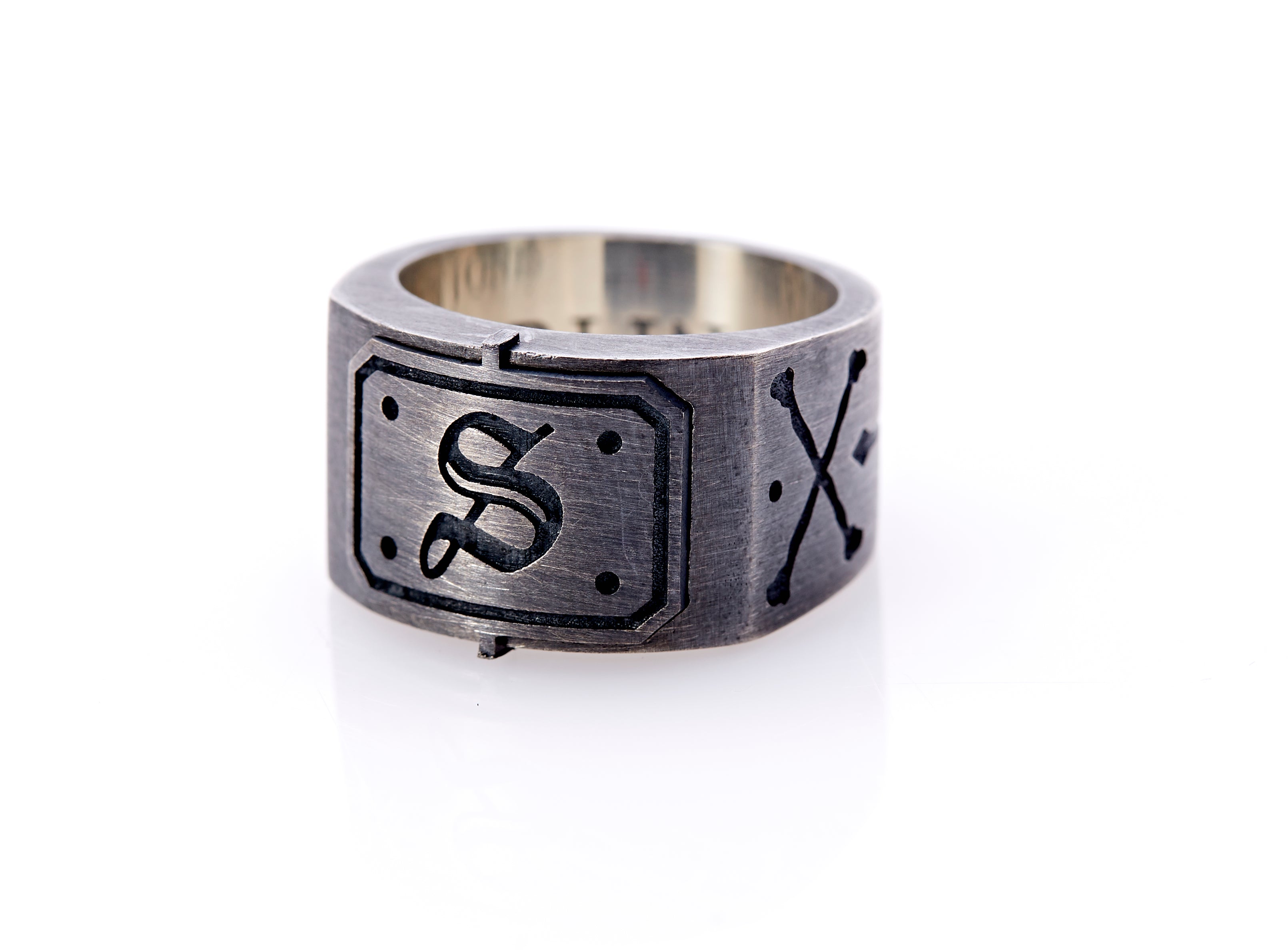 Oxidized sterling silver men’s signet ring with a thick flat band hand-crafted in New York. Semi-raised octagonal engraved top cap with a customizable insignia design of an engraved letter “S” in Old English Style lettering and 4 engraved dots at each corner. The initial on the ring is surrounded by a bold black engraved border that follows the inside edge of the rectangle. The rectangle’s top and bottom edge is accented by a small lip in the center that wraps around the corner edge of the ring.