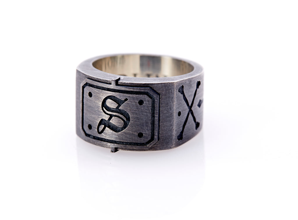 Oxidized sterling silver men’s signet ring with a thick flat band hand-crafted in New York. Semi-raised octagonal engraved top cap with a customizable insignia design of an engraved letter “S” in Old English Style lettering and 4 engraved dots at each corner. The initial on the ring is surrounded by a bold black engraved border that follows the inside edge of the rectangle. The rectangle’s top and bottom edge is accented by a small lip in the center that wraps around the corner edge of the ring.