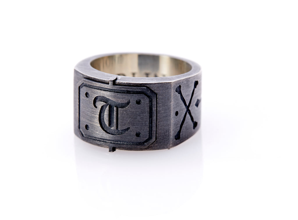 Oxidized sterling silver men’s signet ring with a thick flat band hand-crafted in New York. Semi-raised octagonal engraved top cap with a customizable insignia design of an engraved letter “T” in Old English Style lettering and 4 engraved dots at each corner. The initial on the ring is surrounded by a bold black engraved border that follows the inside edge of the rectangle. The rectangle’s top and bottom edge is accented by a small lip in the center that wraps around the corner edge of the ring.