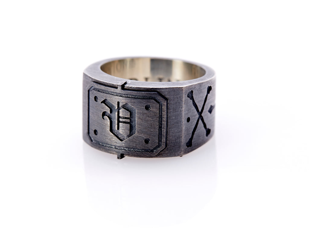 Oxidized sterling silver men’s signet ring with a thick flat band hand-crafted in New York. Semi-raised octagonal engraved top cap with a customizable insignia design of an engraved letter “V” in Old English Style lettering and 4 engraved dots at each corner. The initial on the ring is surrounded by a bold black engraved border that follows the inside edge of the rectangle. The rectangle’s top and bottom edge is accented by a small lip in the center that wraps around the corner edge of the ring.