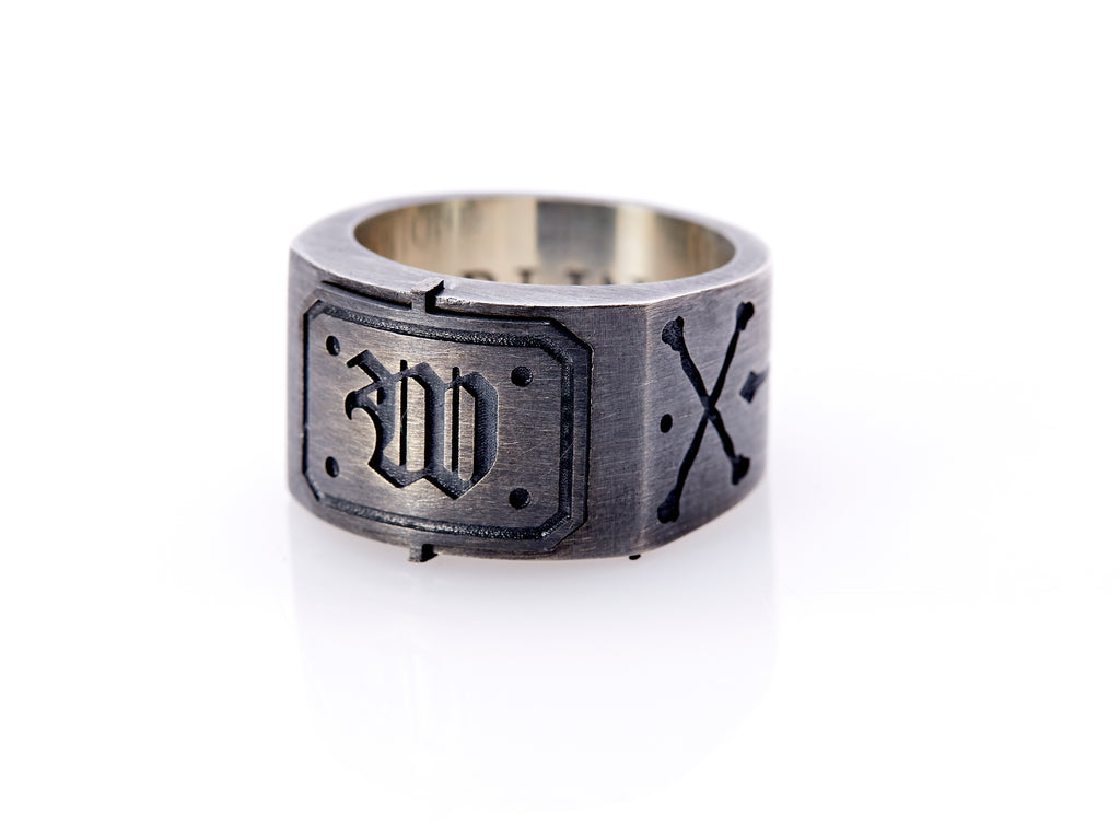 Oxidized sterling silver men’s signet ring with a thick flat band hand-crafted in New York. Semi-raised octagonal engraved top cap with a customizable insignia design of an engraved letter “W” in Old English Style lettering and 4 engraved dots at each corner. The initial on the ring is surrounded by a bold black engraved border that follows the inside edge of the rectangle. The rectangle’s top and bottom edge is accented by a small lip in the center that wraps around the corner edge of the ring.