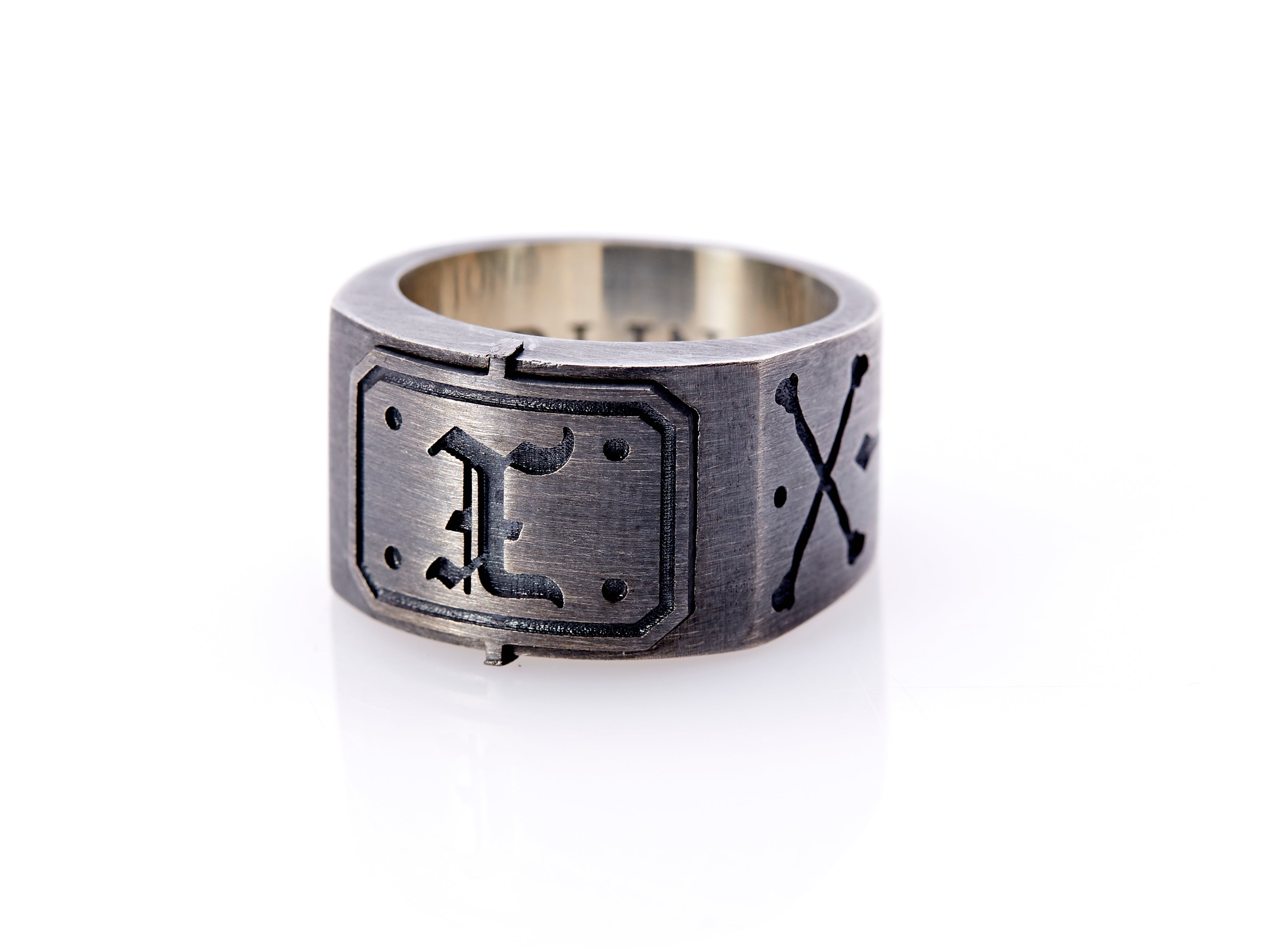 Oxidized sterling silver men’s signet ring with a thick flat band hand-crafted in New York. Semi-raised octagonal engraved top cap with a customizable insignia design of an engraved letter “X” in Old English Style lettering and 4 engraved dots at each corner. The initial on the ring is surrounded by a bold black engraved border that follows the inside edge of the rectangle. The rectangle’s top and bottom edge is accented by a small lip in the center that wraps around the corner edge of the ring.