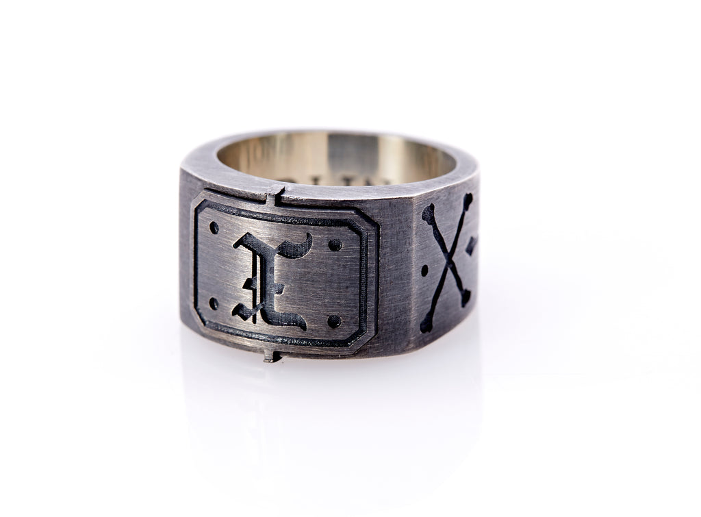 Oxidized sterling silver men’s signet ring with a thick flat band hand-crafted in New York. Semi-raised octagonal engraved top cap with a customizable insignia design of an engraved letter “X” in Old English Style lettering and 4 engraved dots at each corner. The initial on the ring is surrounded by a bold black engraved border that follows the inside edge of the rectangle. The rectangle’s top and bottom edge is accented by a small lip in the center that wraps around the corner edge of the ring.
