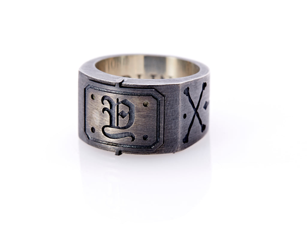 Oxidized sterling silver men’s signet ring with a thick flat band hand-crafted in New York. Semi-raised octagonal engraved top cap with a customizable insignia design of an engraved letter “Y” in Old English Style lettering and 4 engraved dots at each corner. The initial on the ring is surrounded by a bold black engraved border that follows the inside edge of the rectangle. The rectangle’s top and bottom edge is accented by a small lip in the center that wraps around the corner edge of the ring.