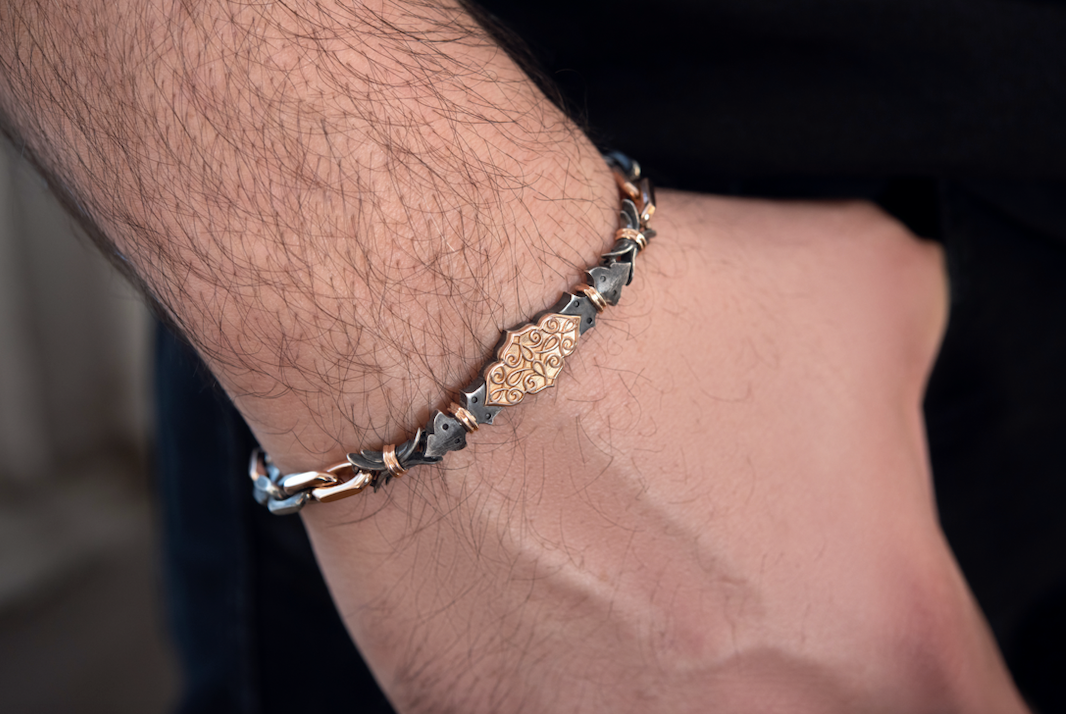 Closeup on man’s wrist wearing the Harlin Jones oxidized sterling silver and rose gold bracelet. The filigree plate sits in the center of the wrist and is roughly the size of a Tic Tac. As the arm points downwards, the bracelet falls to where the wrist meets the hand. The bracelet is the width of a thick rubber band.