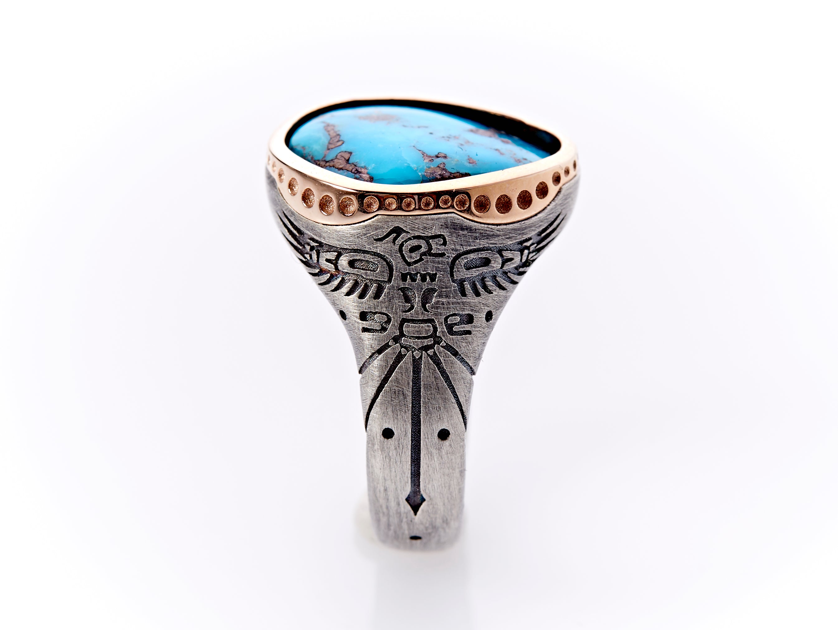The sterling silver ring band has a hand-engraving of a tribal Thunderbird design accented with 5 dots, 2 flanking the body, 2 flanking a long tail feather with an arrow point extending down the center of the band, and 1 below the arrow point. The wings of the Thunderbird look as if they are supporting the rose gold bezel setting, which has custom circular engravings of varying sizes. A unique shape of turquoise with iron pyrite flakes is handset in the rose gold setting.