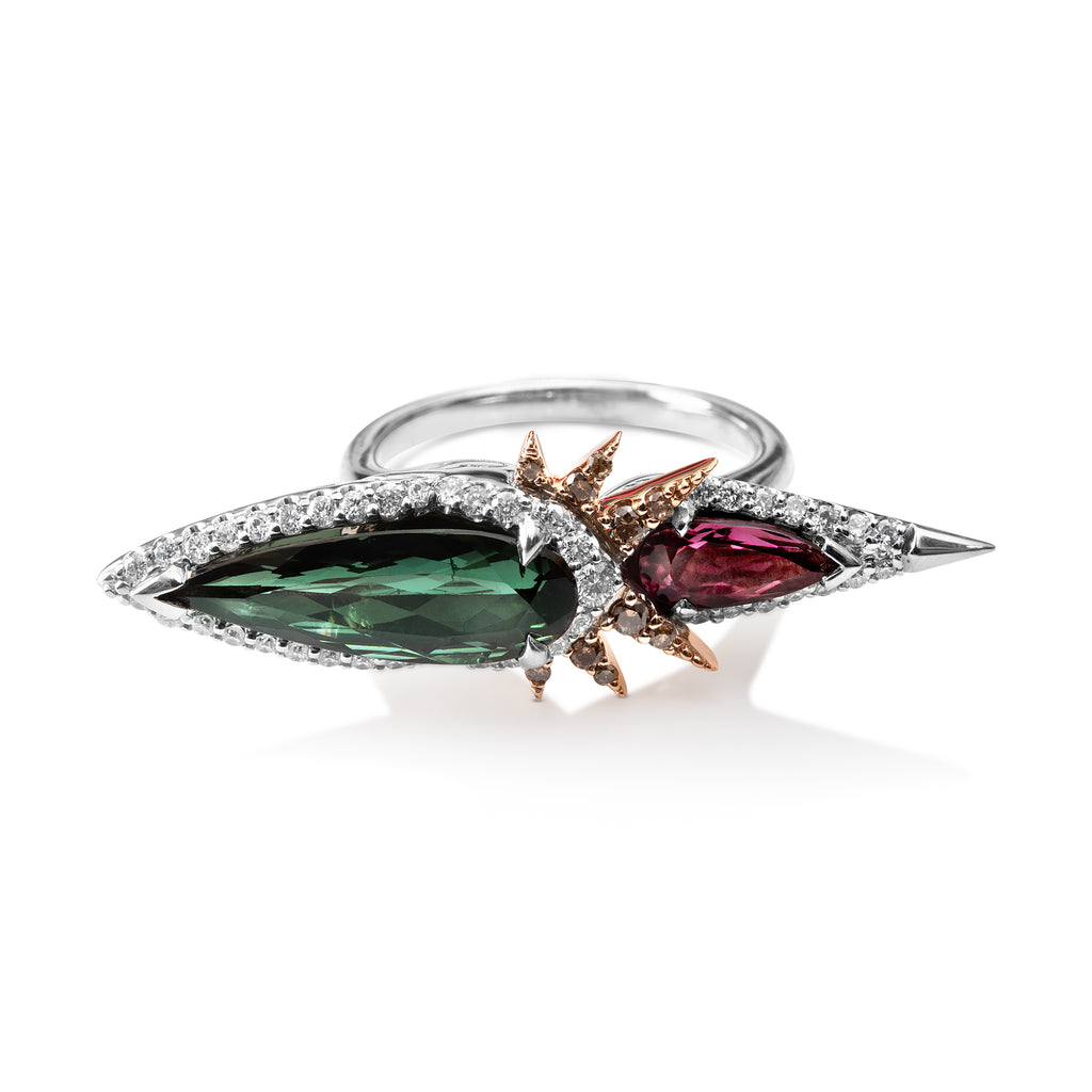 Bespoke tourmaline and diamond ring with thin, round white gold band. Pear- shaped green tourmaline gem surrounded by round diamonds. The pointed edge extends horizontally from the ring’s center. A smaller pink tourmaline pear-shaped gem extends horizontally in the opposite direction, surrounded by round diamonds. The pink stone has a sharp white gold point at the end and is set below the green gem. 6 rose gold diamond spikes extend out from center to overlap the pink gem.