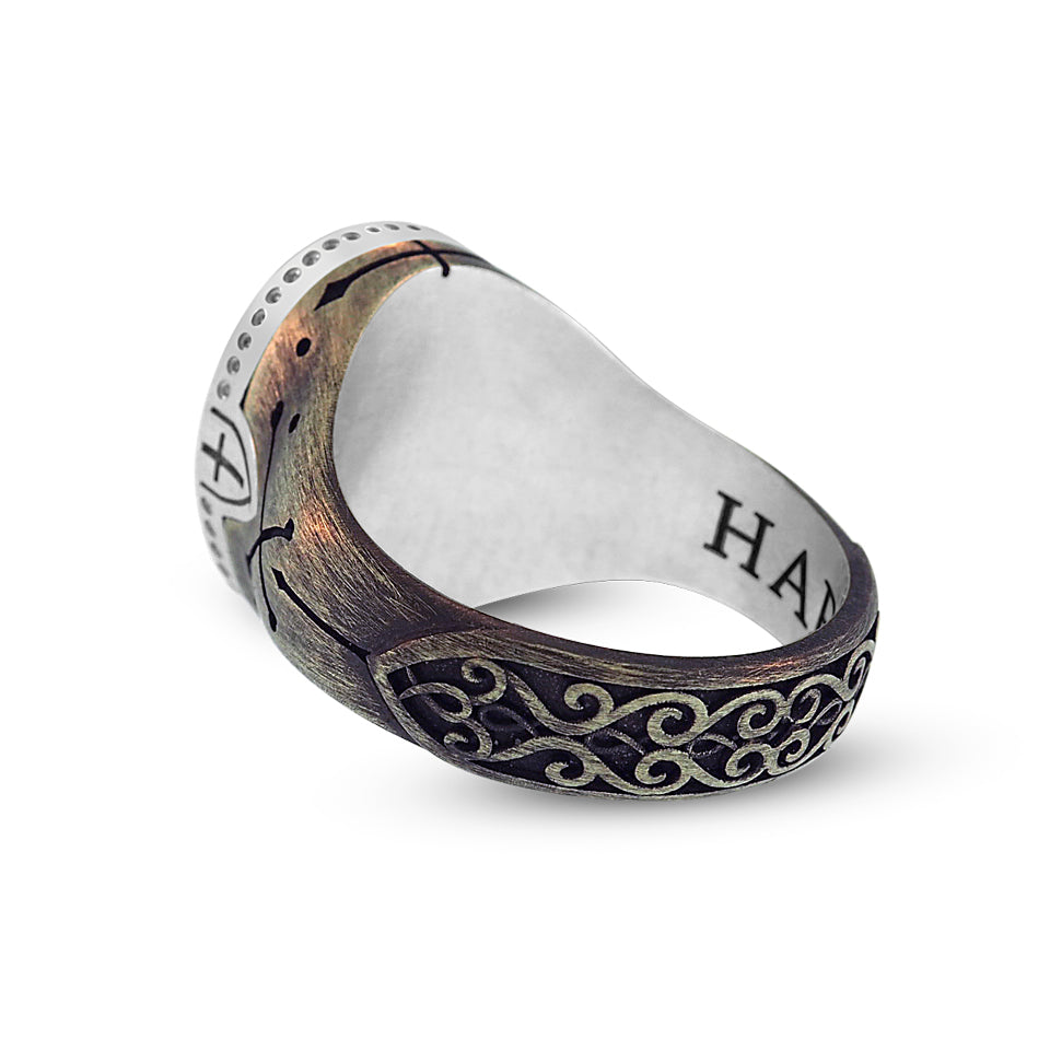 Partial back view of the Turquoise Cross-Bones Ring. An ornate relief of decorative interlocking swirls against a dark background extends from the base of the ring to the lower shank. The filigree pattern has a thick border and tapers into a point that leads to a hand engraved arrow that extends up the side of the ring.