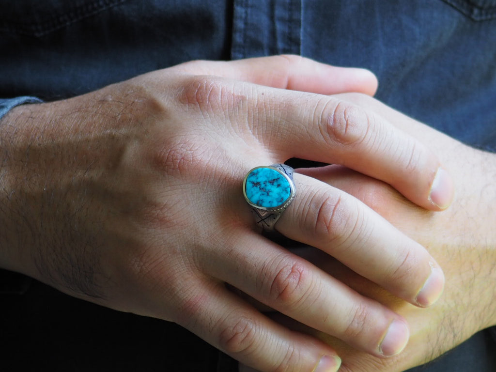 Closeup on man’s hand wearing the Turquoise Cross-Bones Ring on his middle finger. The ring’s band fits low around the base of the knuckle. The ring’s stone extends from the base of the knuckle to halfway below the first joint, with the cross-bones on the side of the band visible.