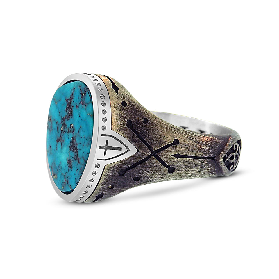 Partial side view of the men’s turquoise ring shows the large stone setting tapering into a thin metal band. Polished sterling silver bezel setting sits like a cap on the ring with a continuous pattern of small circular engravings around the perimeter. There is an engraving of a cross within a shield on the bezel on either side of the stone that drops into the band. The sterling silver band has a custom engraving of cross-bones that sit just below the cross and shield.