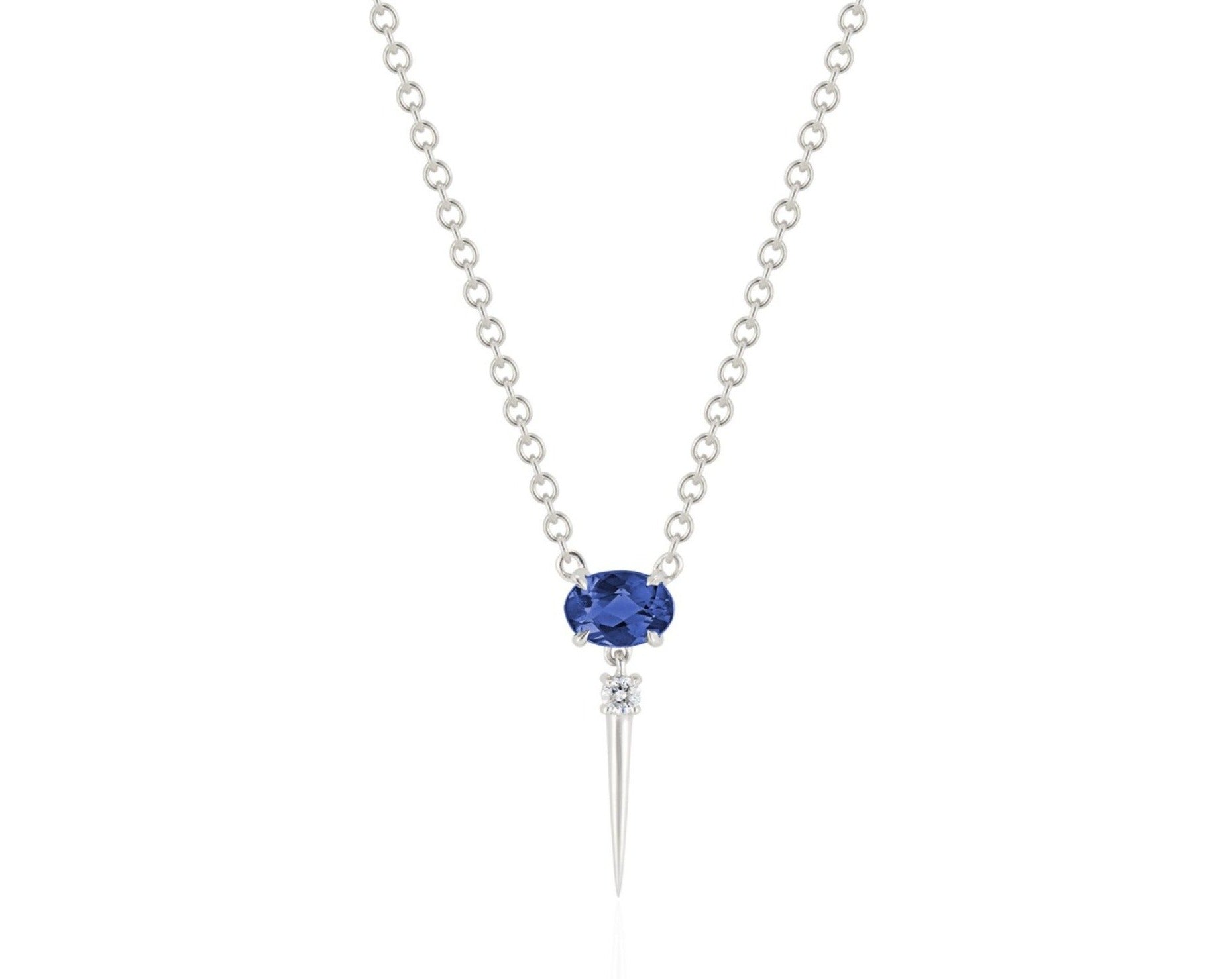 Front view of blue sapphire drop necklace. A horizontal oval-shaped blue sapphire is fixed with 4 prongs to a white gold round-link chain. Below this, a single link connects to a small round diamond with a dramatic white gold spike dropping down.