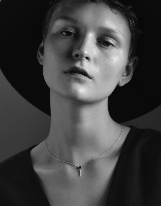 A black-and-white image of a person wearing a hat and a white gold necklace with a blue sapphire pendant.