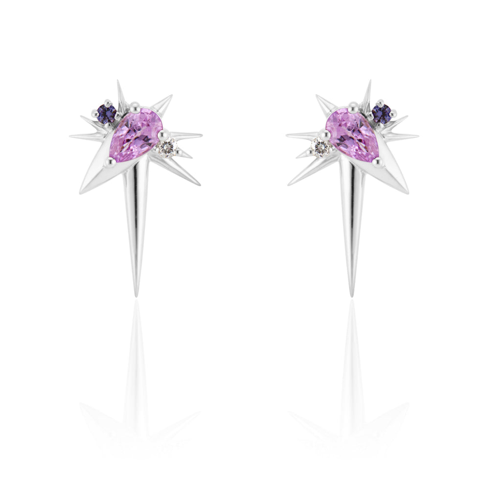  Front view of slanted drop earrings, each with 1 pear-shaped pink sapphire, set at 45-degree angle, flanked by small round diamonds and sapphires. 9 white gold spikes of varying lengths extend from the oval setting, with a larger and longer spike pointed down.
