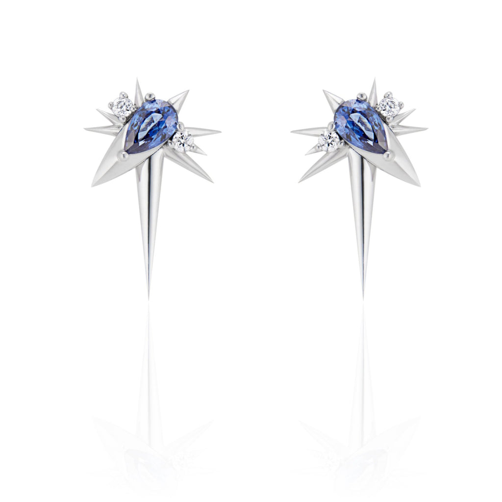 Front view of slanted drop earrings, each with 1 pear-shaped blue sapphire, set at 45-degree angle, flanked by small round diamonds. 9 white gold spikes of varying lengths extend from the oval setting, with a larger and longer spike pointed down.