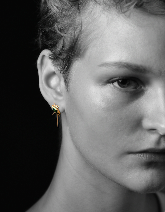 Close-up of an ear with a dragonfly earring against a black background; part of the image is obscured.